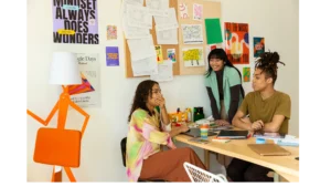 Three young creatives brainstorming fashion design ideas at a desk, with sketches and colorful posters on the wall behind them.